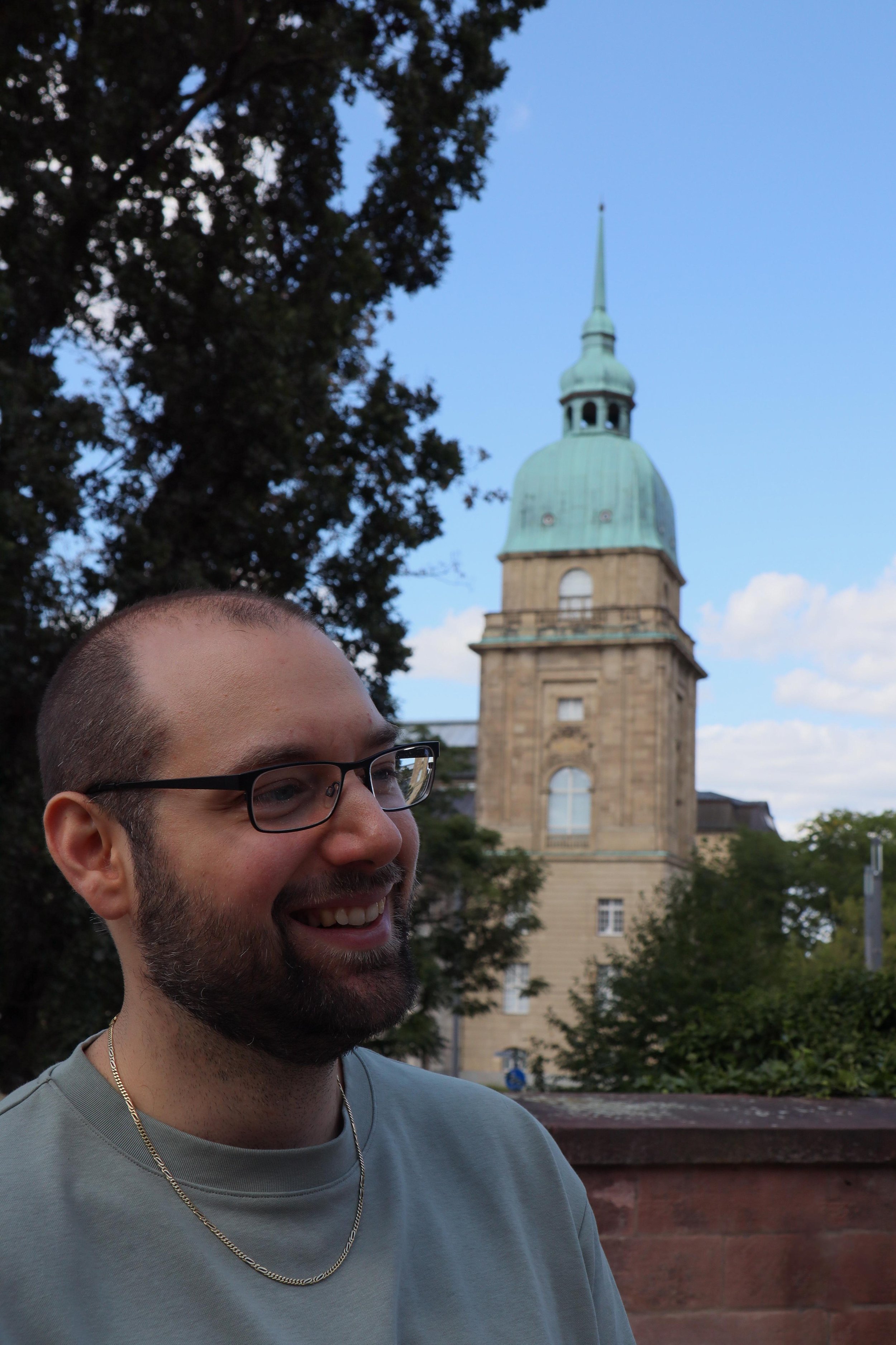 Levent Wachtl im Zentrum Darmstadts vor einem historischen Gebäude mit grünem Turm, umgeben von Bäumen und Blauen Himmel.