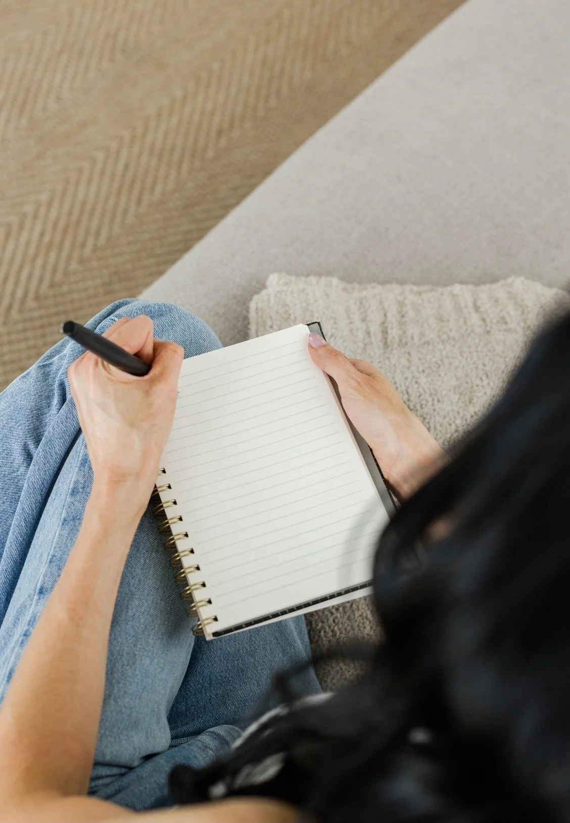A person sitting on a chair or couch, wearing blue jeans and holding a black marker, preparing to write in an open, lined notebook resting on their lap, with a textured rug and carpeted floor visible in the background.