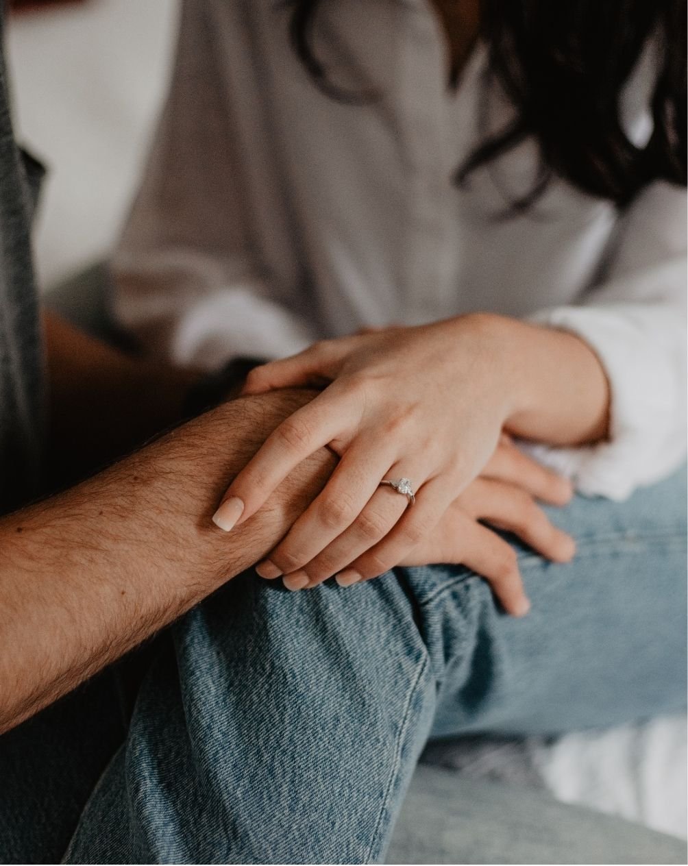 Close-up of a woman's hand with an engagement ring resting on a man's hand, with both hands resting on her knee, in a casual setting.