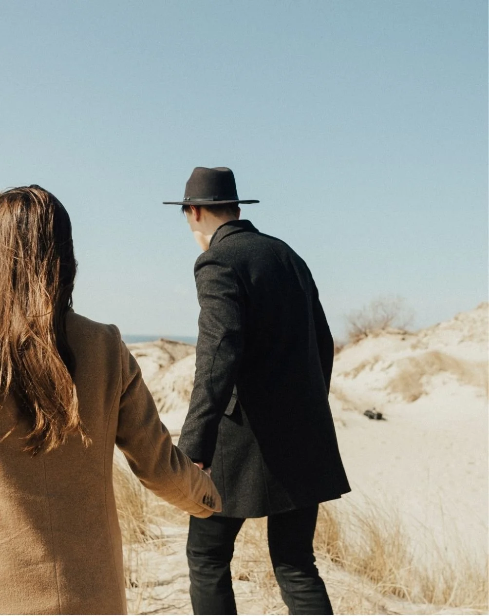 A man and woman holding hands walking along a sandy beach or dunes under a clear blue sky. The man is wearing a dark long coat and a wide-brimmed hat; the woman is wearing a tan coat.