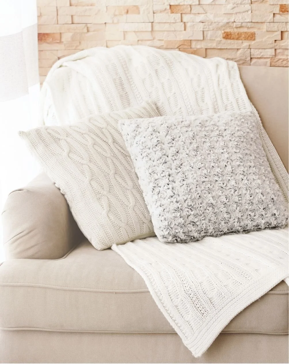 A cozy beige sofa decorated with knitted and furry white pillows and a white textured blanket, against a brick wall background.