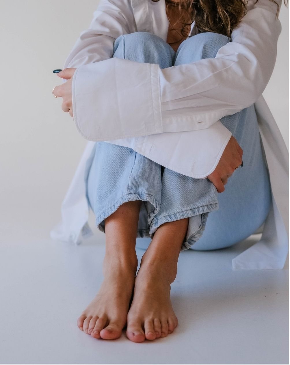 A person sitting cross-legged on the floor, wearing a white shirt and light blue jeans, with their arms wrapped around their knees.