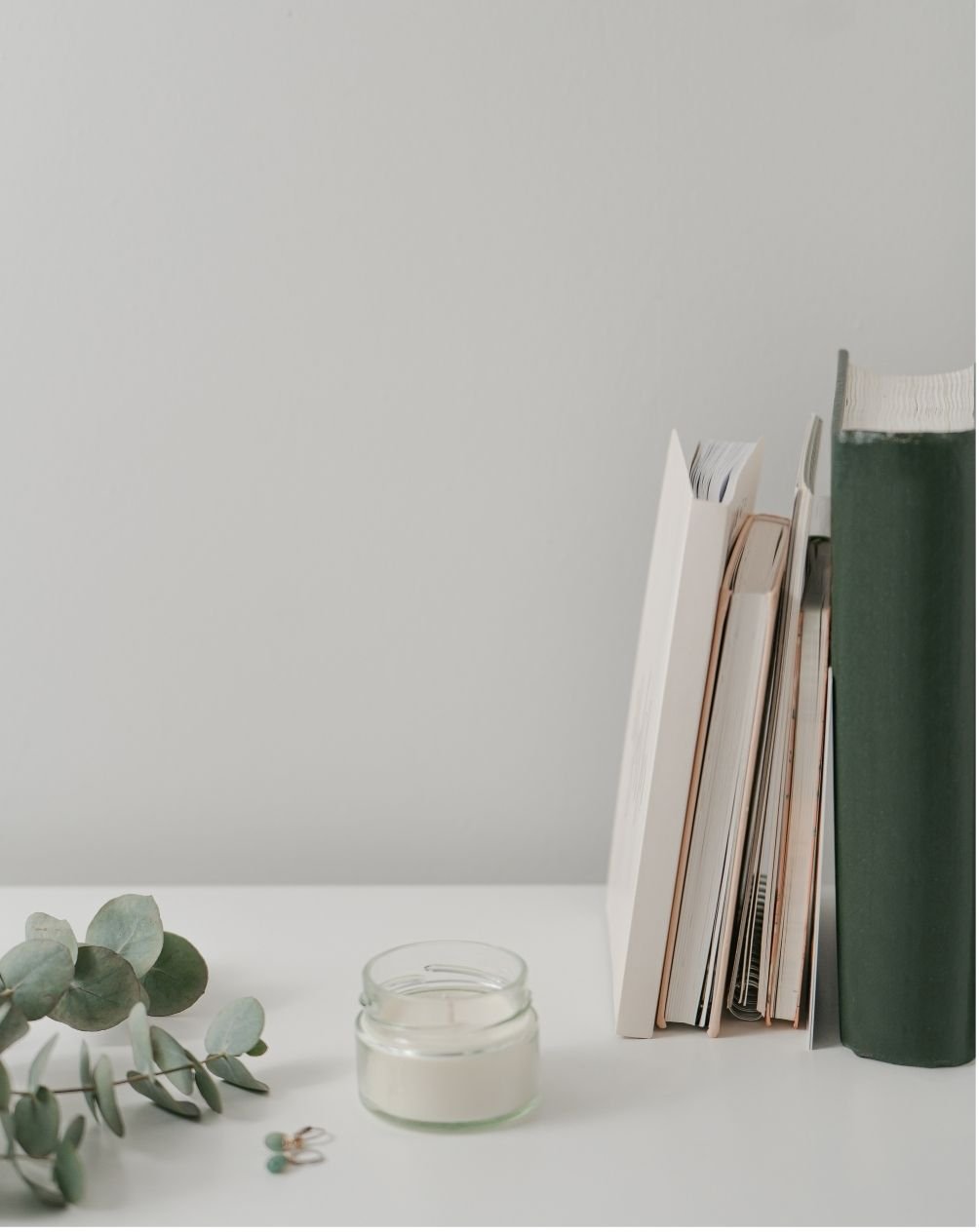 Books, a jar of white candle, and eucalyptus leaves on a white surface.