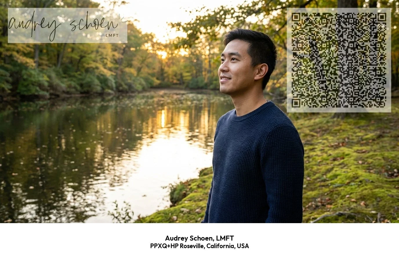 A man stands on the edge of a river during sunset, surrounded by trees with autumn foliage, wearing a dark blue sweater, and smiling as he looks into the distance.
