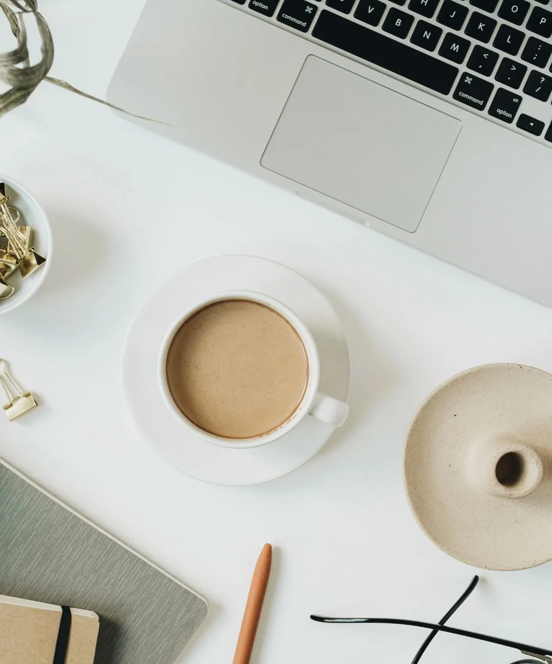 A white desk with a laptop, a cup of coffee on a saucer, a notebook, a pen, a small bowl filled with gold binder clips, and a beige decorative object placed on it.