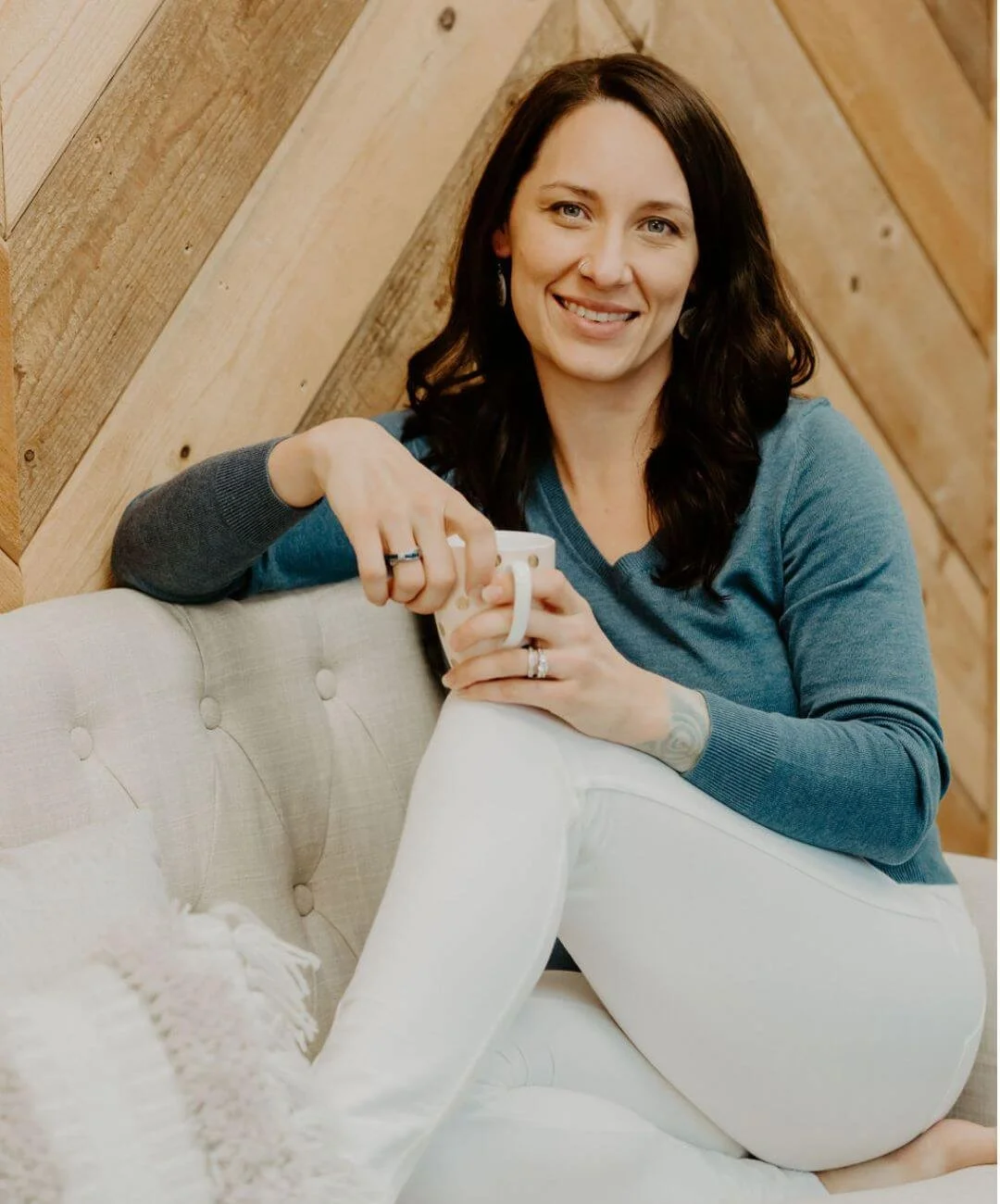 Audrey Schoen, therapist in Roseville, CA, with dark brown hair, wearing a blue long-sleeve shirt and white pants, sitting on a beige tufted sofa, holding a white mug, in front of a wooden wall background, smiling at the camera.