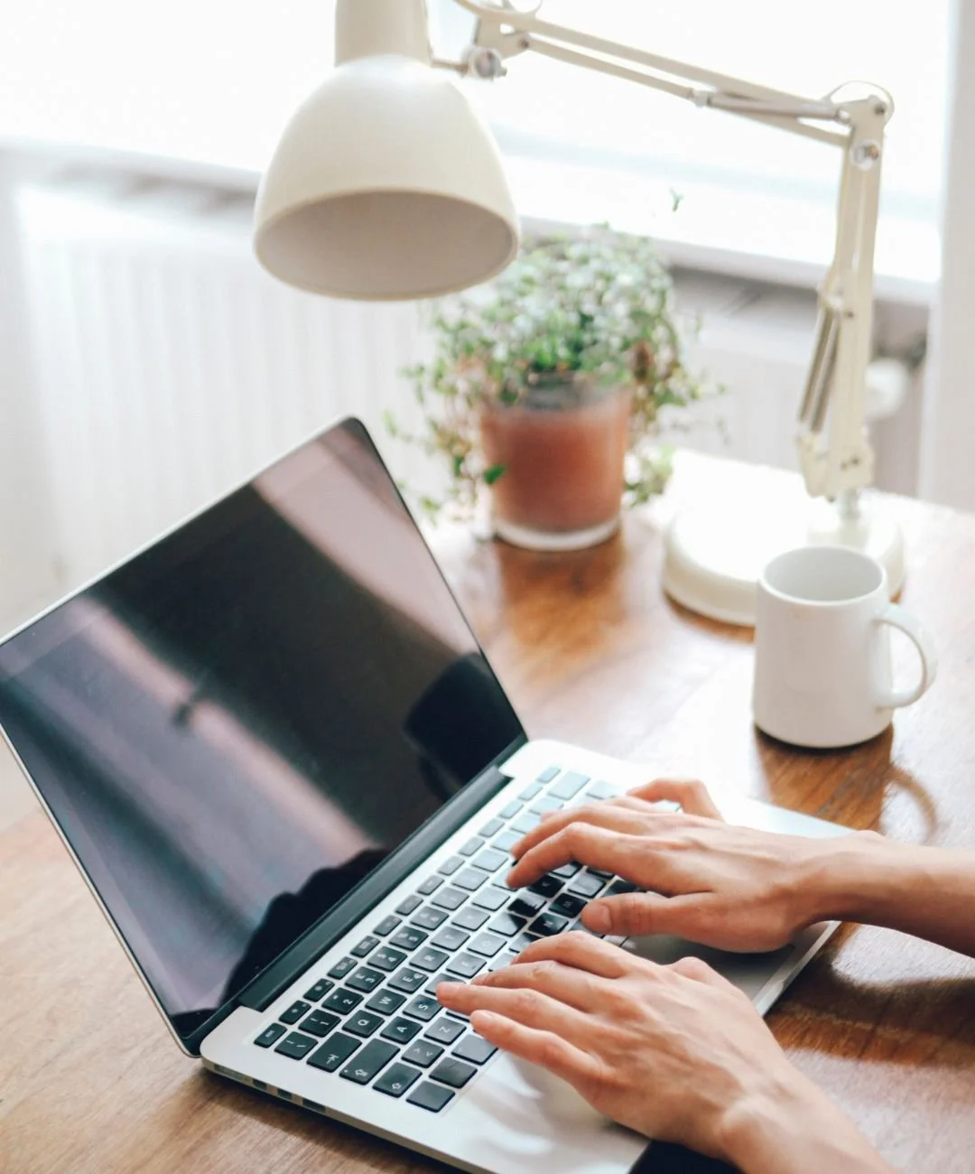 Person typing on a laptop at a wooden desk with a potted plant, white mug, and desk lamp, near a window.