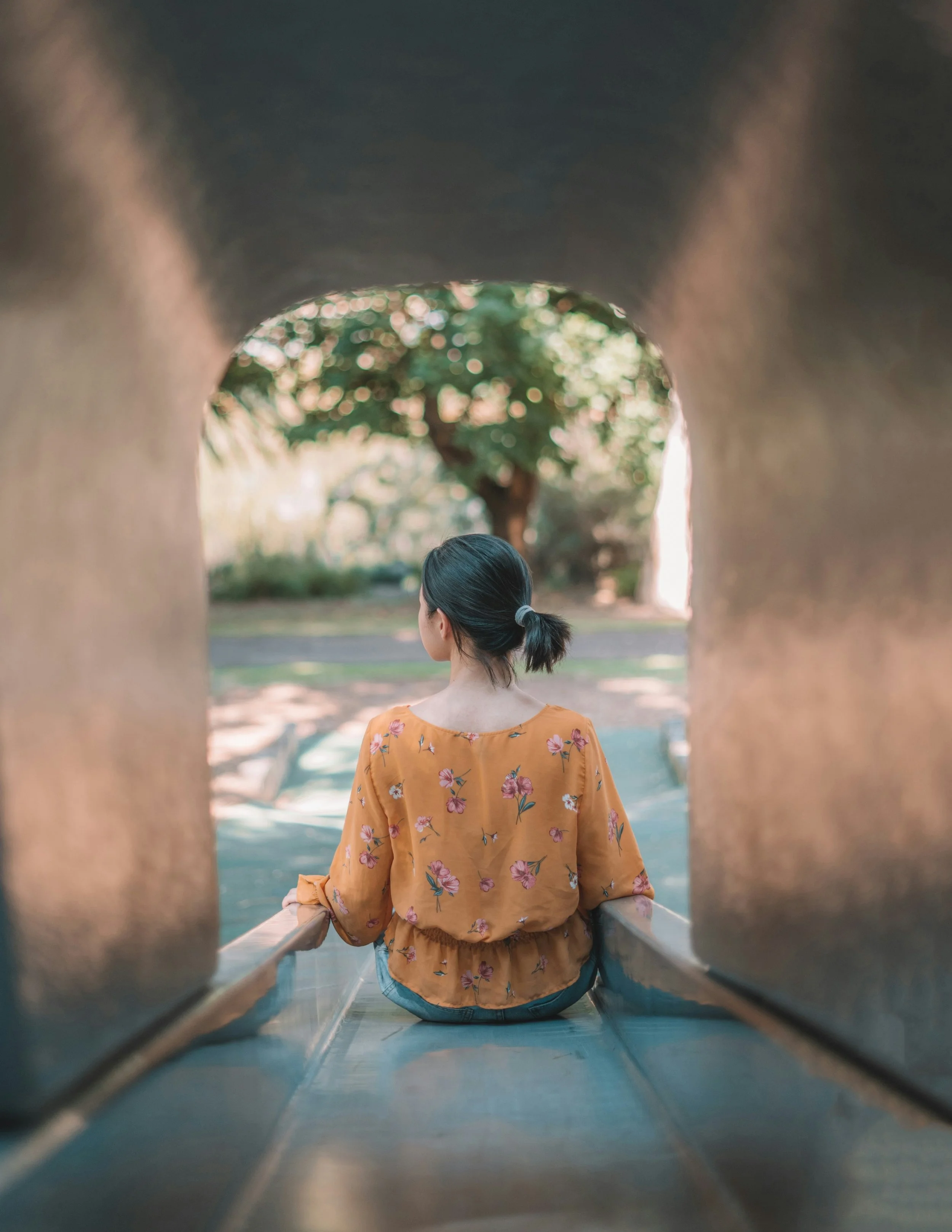 A woman with dark hair tied in a bun, wearing a yellow floral top, sits on a slide at a park, facing away and looking toward trees and a park path.