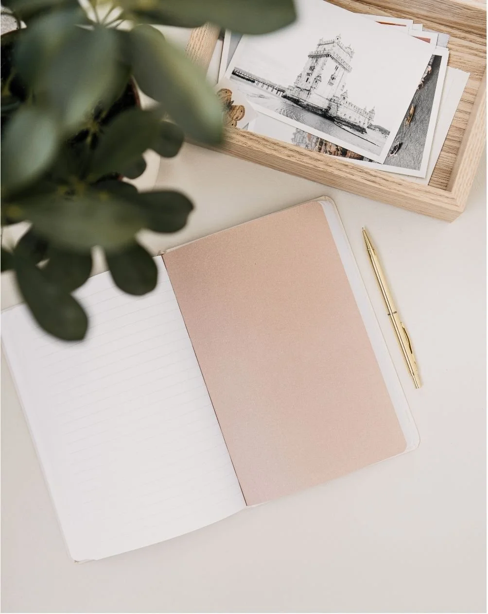 Open notebook with lined pages and a pink section, gold pen on the right, wooden tray with black and white photographs, green plant partially visible, light-colored surface.