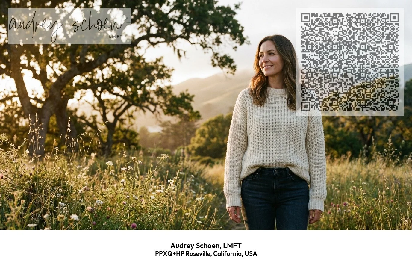 A woman with long brown hair, wearing a cream sweater and dark jeans, standing in a field of wildflowers with trees and hills in the background, smiling and looking to the side during sunset.