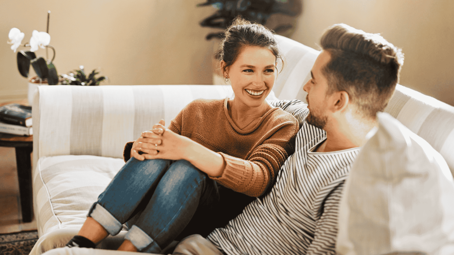A young woman and man sitting on a couch and smiling at each other, engaging in conversation in a cozy living room.