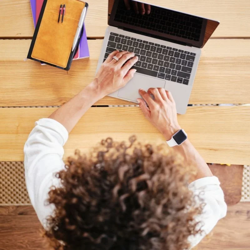 A person with curly hair working on a laptop at a wooden desk, surrounded by notebooks and pens.