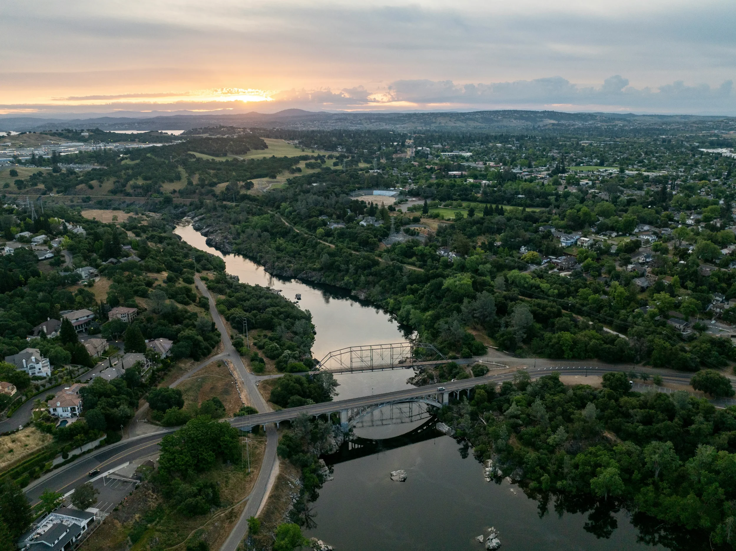 Aerial view of a city landscape during sunset with a river running through green hills, residential neighborhoods, and scattered buildings, with mountains in the background.