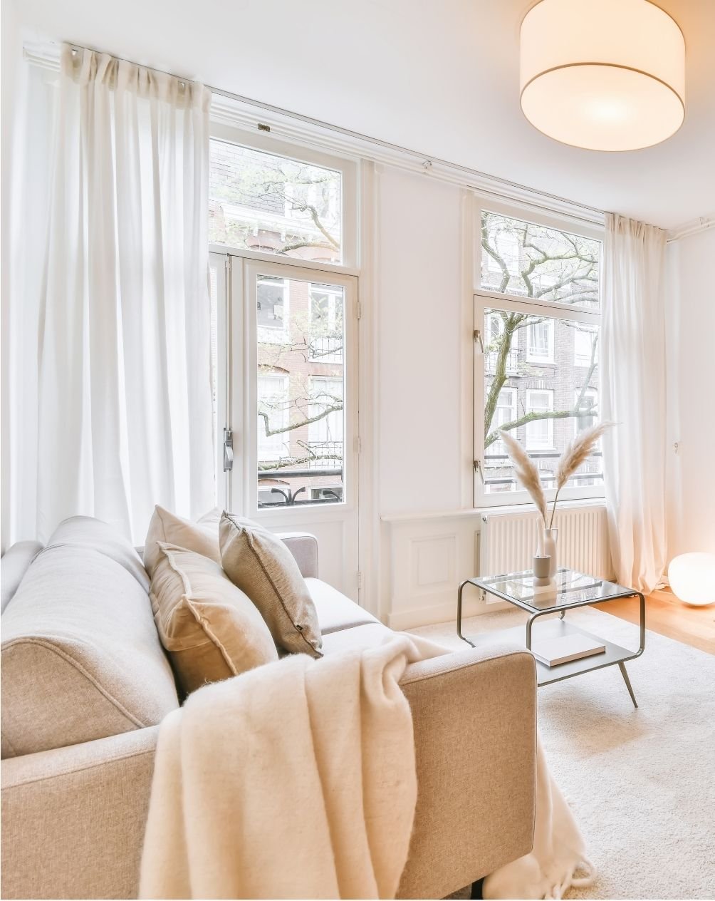 Bright living room with large windows, white curtains, beige sofa with pillows, glass coffee table with a vase of pampas grass, and soft lighting.