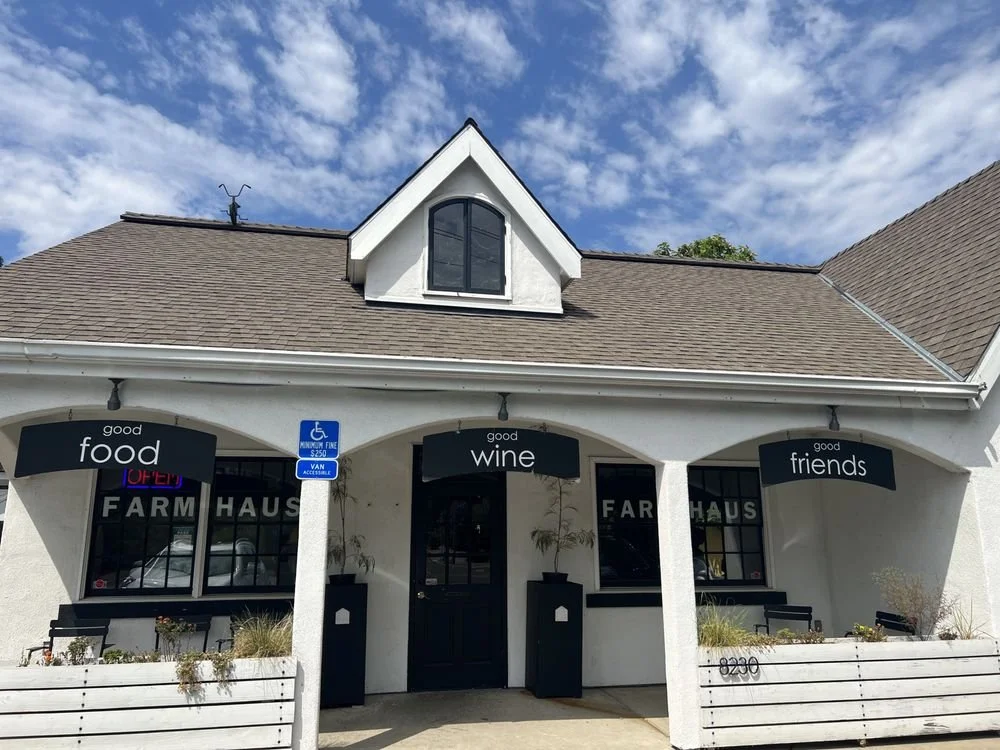 Front view of a farm-to-table restaurant named Farmhaus with an outdoor seating area, three black signs reading "good food," "good wine," and "good friends," and a blue handicap parking sign, under a partly cloudy sky.