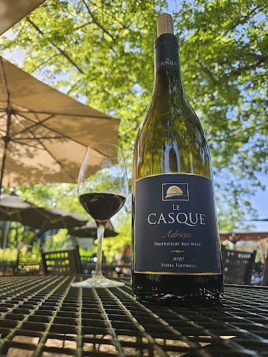 A bottle of Le Casque Adriatic Proprietary Red Wine next to a glass of red wine on a black metal table outdoors with green trees and a patio umbrella in the background.