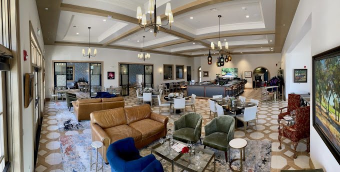 Spacious hotel lounge with high ceilings, multiple seating areas with chairs and sofas, chandeliers, flat-screen TVs, and a bar counter.