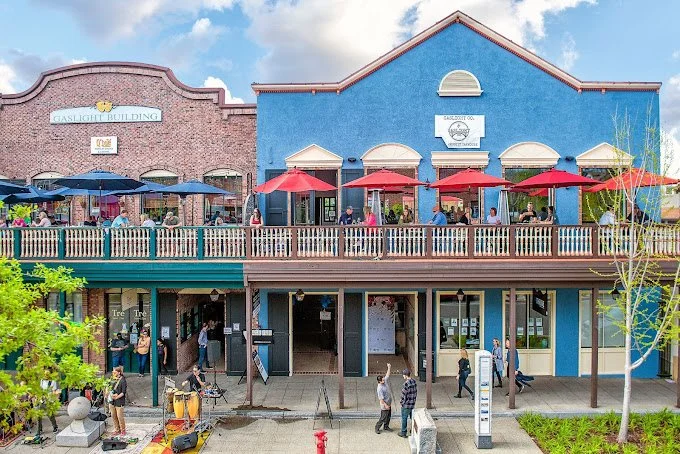 A lively street scene with a two-story building housing cafes and shops, outdoor seating with red umbrellas on the upper level, and people walking on the sidewalk.