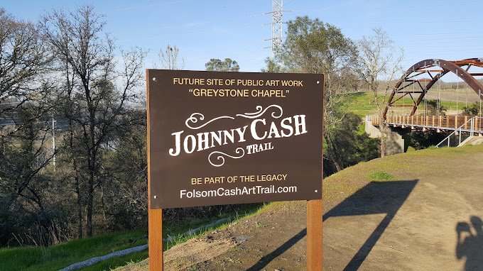 Sign for the Johnny Cash Trail indicating future site of public art work 'Greystone Chapel' and website FolsomCashArtTrail.com, with a bridge and trees in the background.