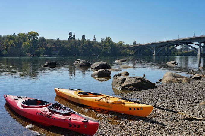 Two kayaks, one red and one yellow, are on the shore of a calm river with large rocks. In the background, there is a bridge and trees lining the riverbank under a clear blue sky.