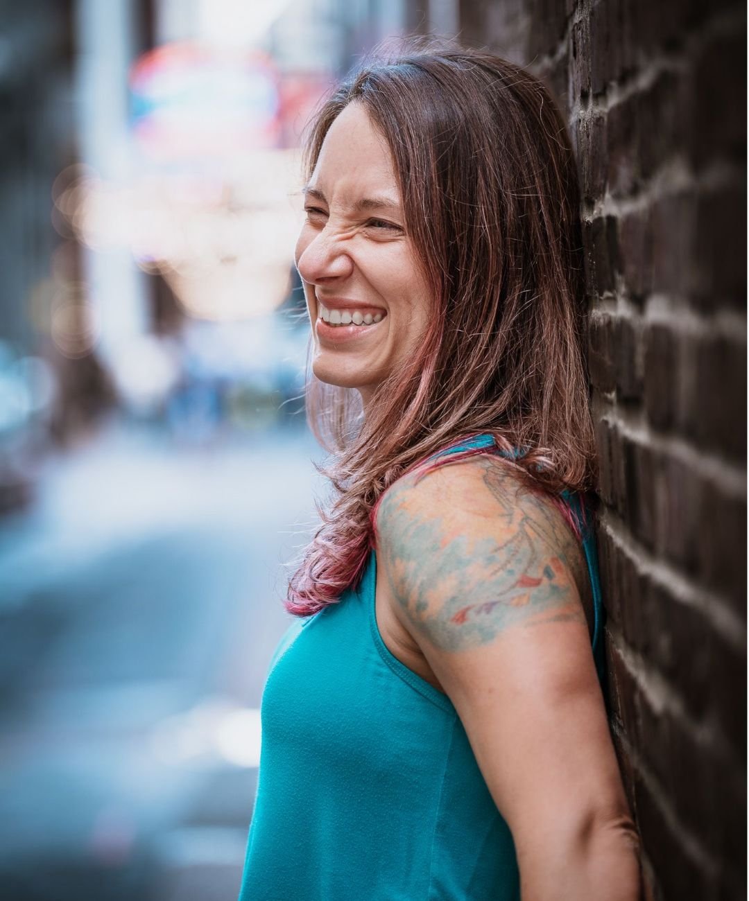 A woman with long brown hair and a colorful tattoo on her shoulder, wearing a teal sleeveless top, leaning against a brick wall and smiling.