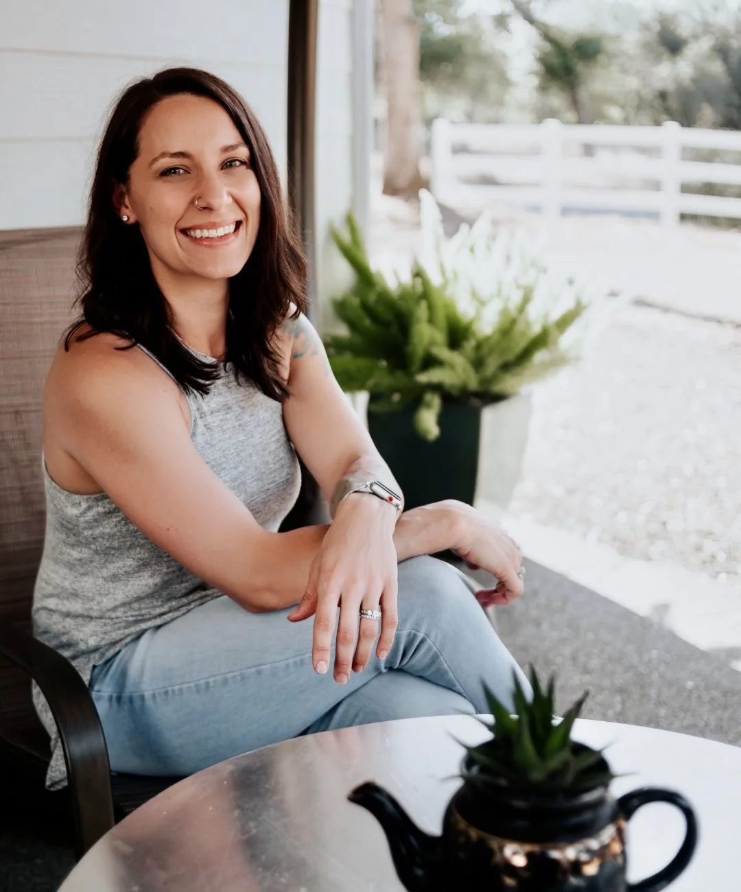Audrey Schoen, therapist in Roseville, CA, with dark brown hair, smiling, sitting at a table outside, wearing a grey top and light blue jeans, with a cactus-shaped plant teapot on the table, and a large green plant in a pot behind her.