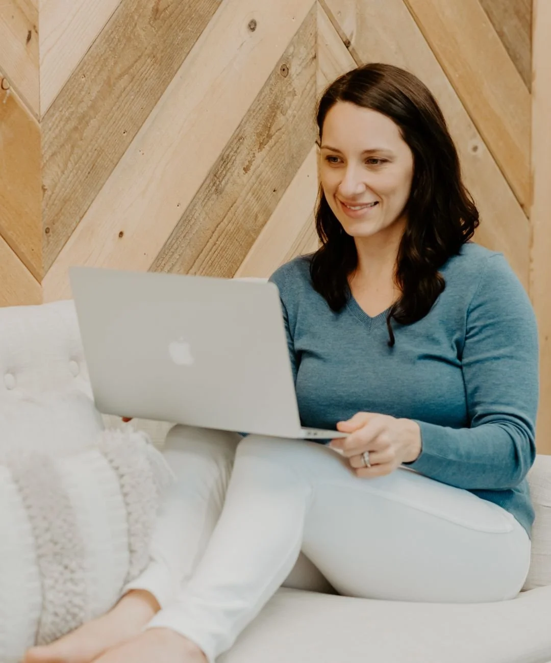 Audrey Schoen, therapist in Roseville, CA, with dark brown hair smiling while using a silver MacBook laptop, sitting on a white couch with a wooden wall background.
