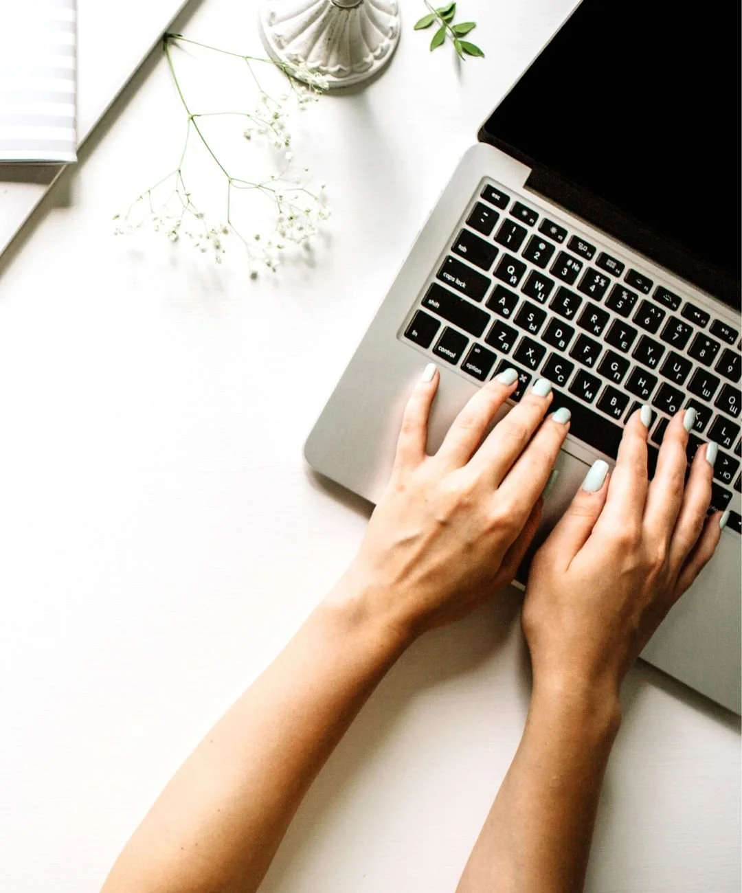 Person typing on a laptop keyboard on a white desk, with a plant, notebook, and a decorative vase nearby.