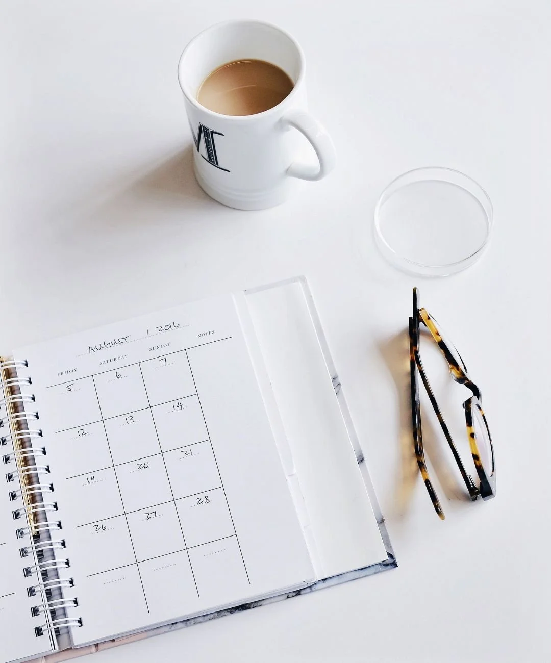 A white desk with a calendar open to August 2016, a pair of tortoiseshell glasses, a small circular container, a pen, and a mug with coffee.