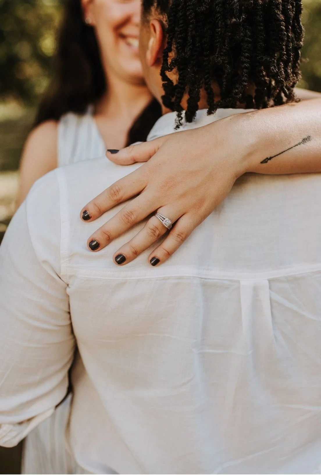 Close-up of a woman with black nail polish showing her hand resting on a man's shoulder. The woman has a wedding ring on her ring finger and a small arrow tattoo on her inner arm. Both are smiling and outdoors with blurred greenery in the background.