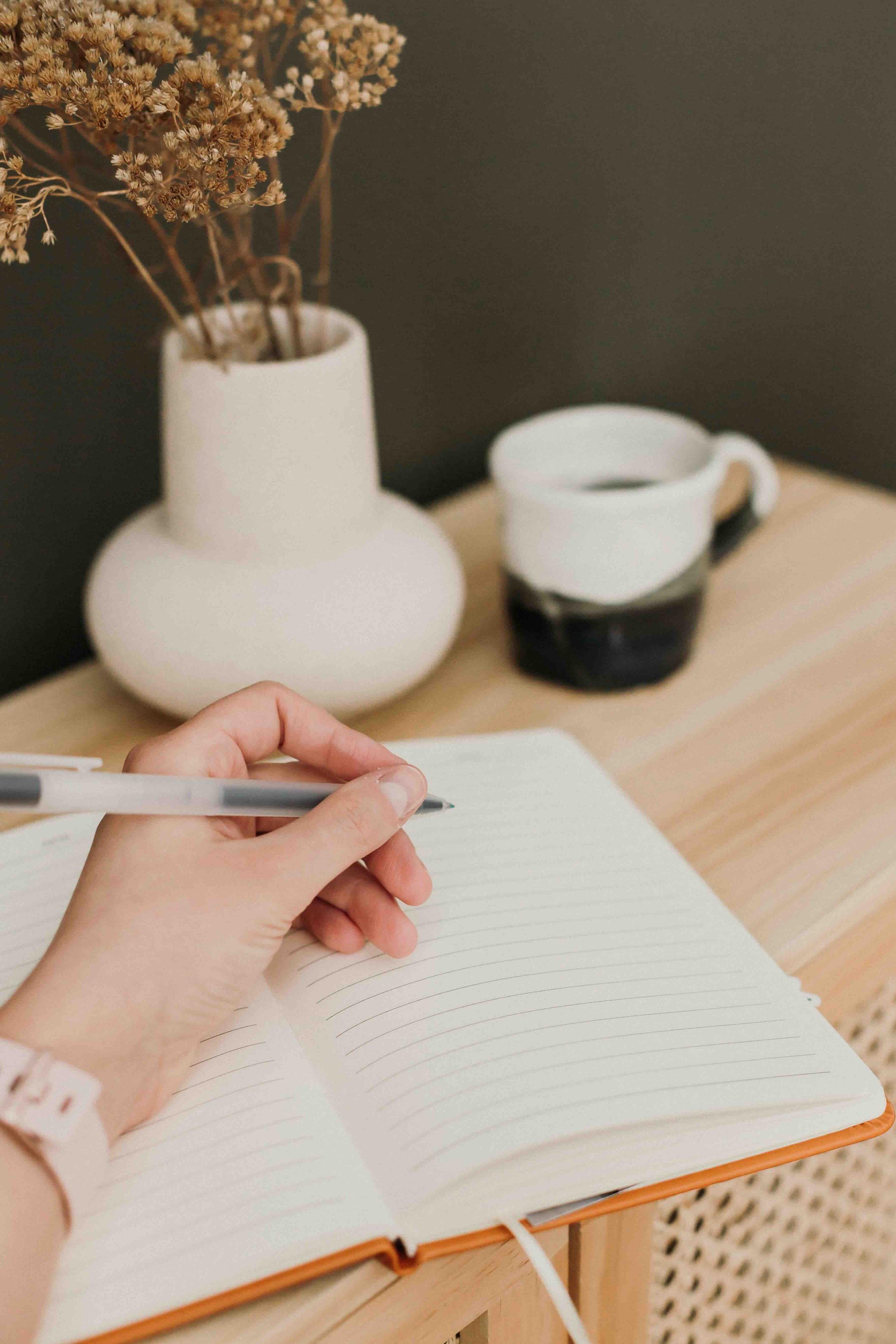 A hand writing in a lined notebook with a black pen. In the background, there is a white vase with dried flowers and a white cup filled with a dark liquid, placed on a wooden surface.