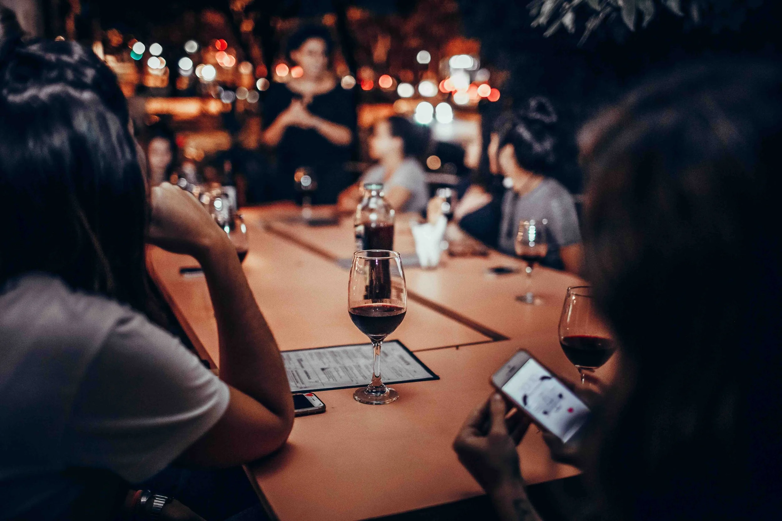 People sitting at a table on a patio at night, drinking wine, using their phones, with a blurry background of city lights.
