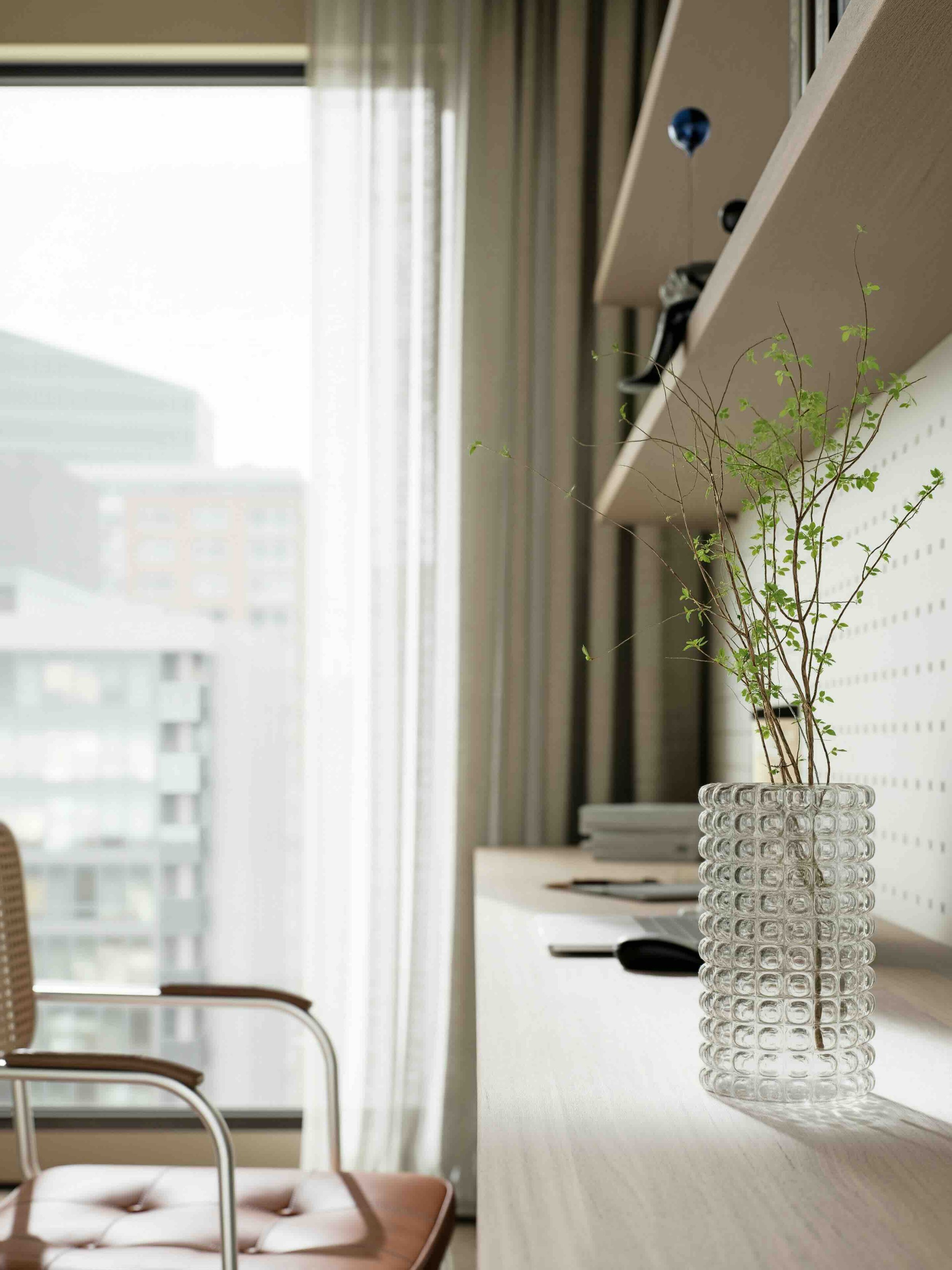 A white, textured glass vase with a branch of small green leaves placed on a white table in an office or living room with tall windows and curtains in the background.