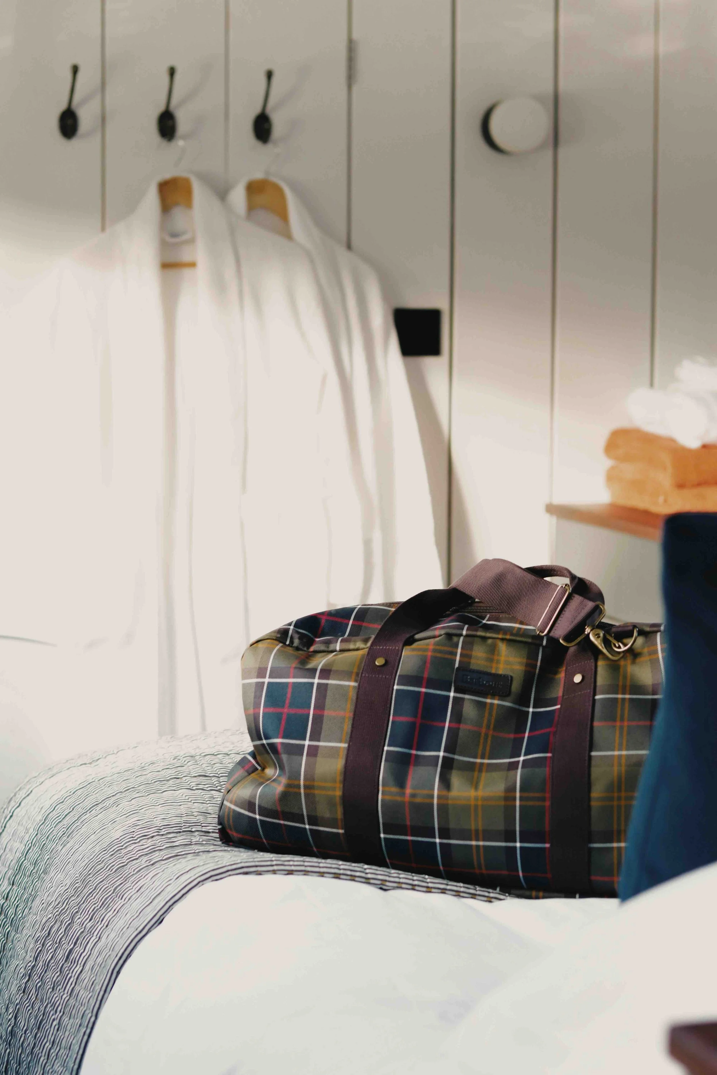 A plaid duffle bag with leather straps and handles placed on a bed with a black and white striped blanket, in a room with white panel walls, closets, and a small side table.