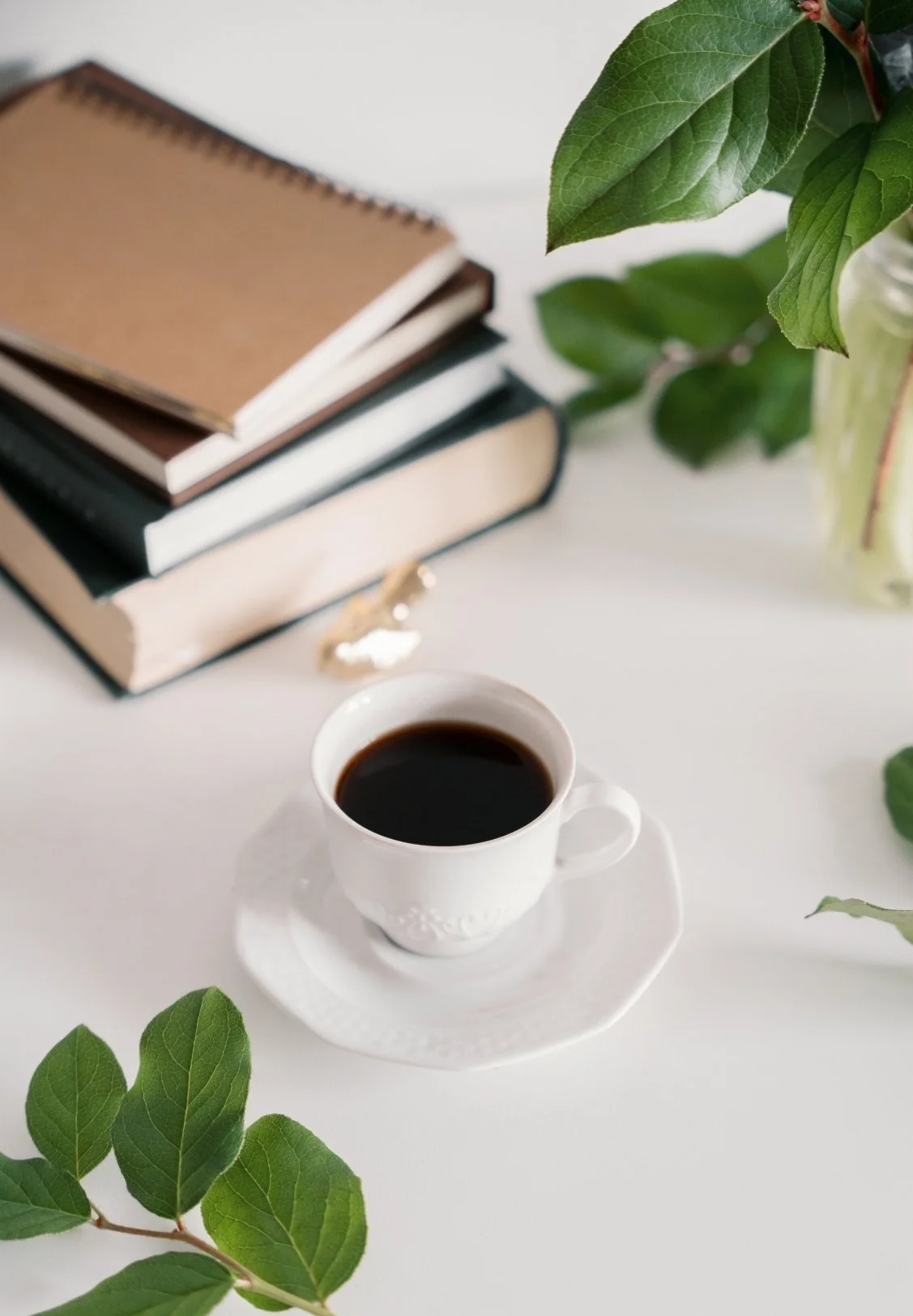 A white cup filled with black coffee on a matching saucer, placed on a white surface. To the left of the cup, there is a branch with green leaves. In the background, there are stacked notebooks with brown, black, and white covers, and a glass jar filled with green leaves and stems.