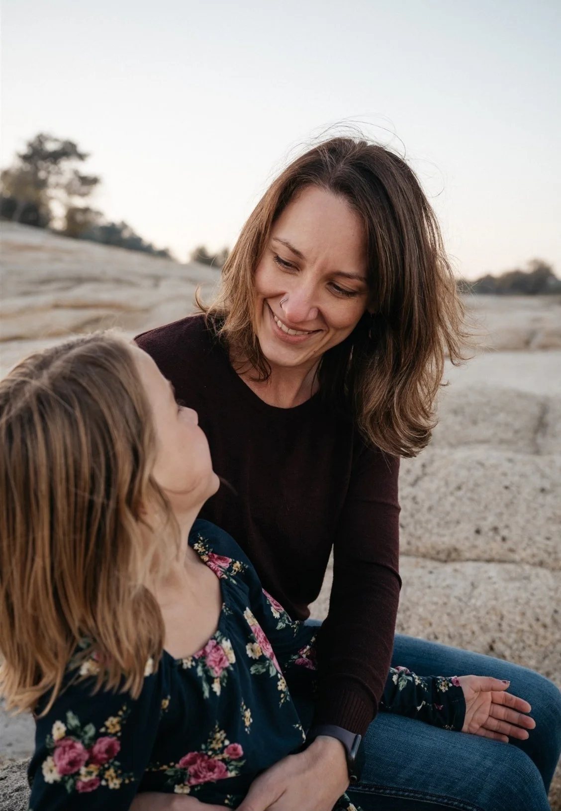 A woman and a young girl sit on a rocky outdoors, looking at each other and smiling.