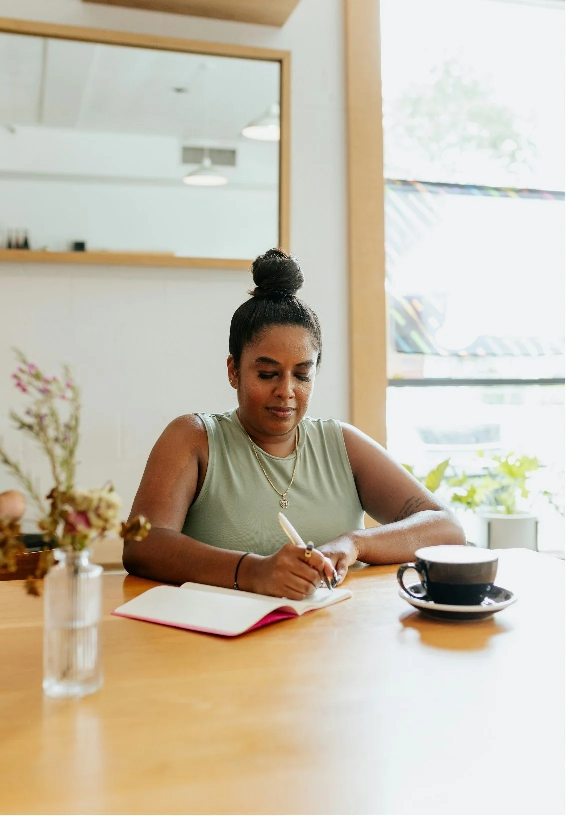 A woman sitting at a wooden table, writing in a notebook with a pen. There are flowers in a vase, a black coffee cup, and a saucer on the table, with sunlight coming through a large window behind her.