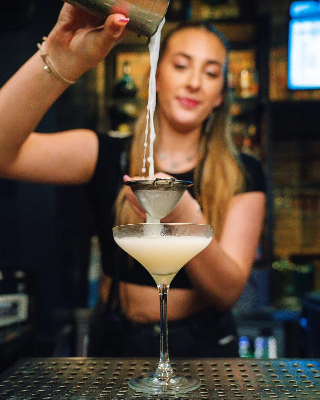 A woman pouring a cocktail through a strainer into a martini glass in a bar.