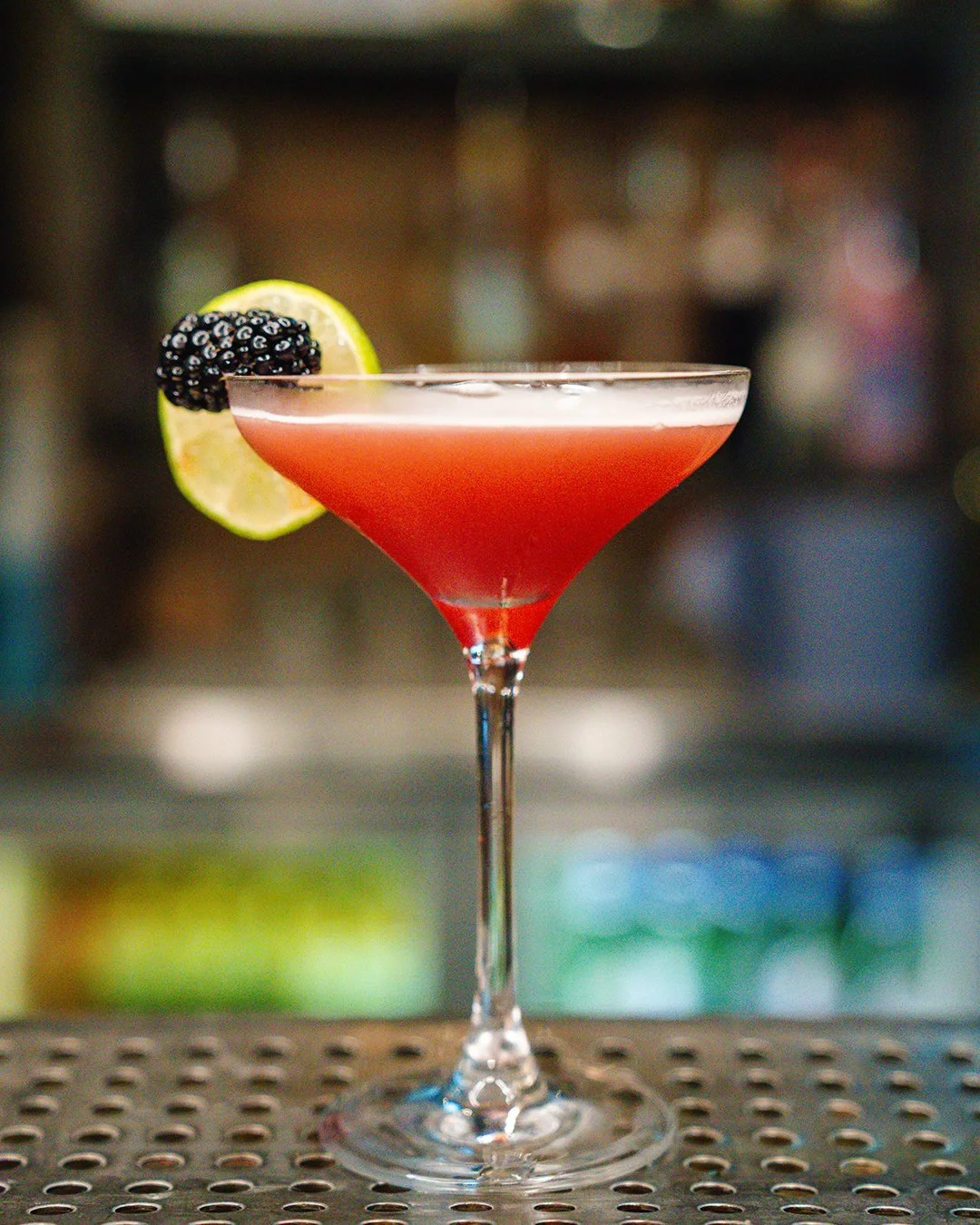 A pink cocktail in a martini glass garnished with a lime wedge and blackberries, placed on a perforated bar surface with a blurred bar background.