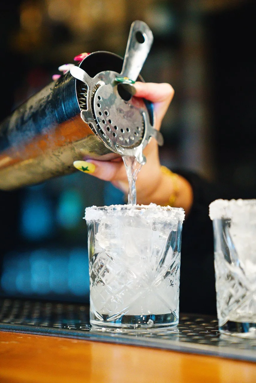 A person is pouring a clear liquid, likely a cocktail or liquor, from a cocktail shaker into a glass rimmed with salt or sugar, on a bar counter.