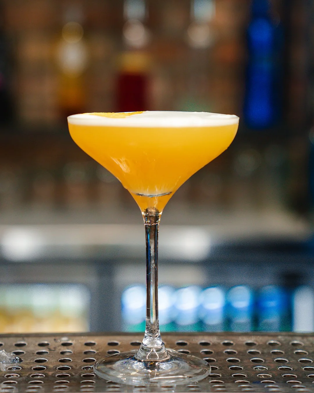 A cocktail in a coupe glass with a yellow-orange color and a lemon garnish on top, set on a bar counter.