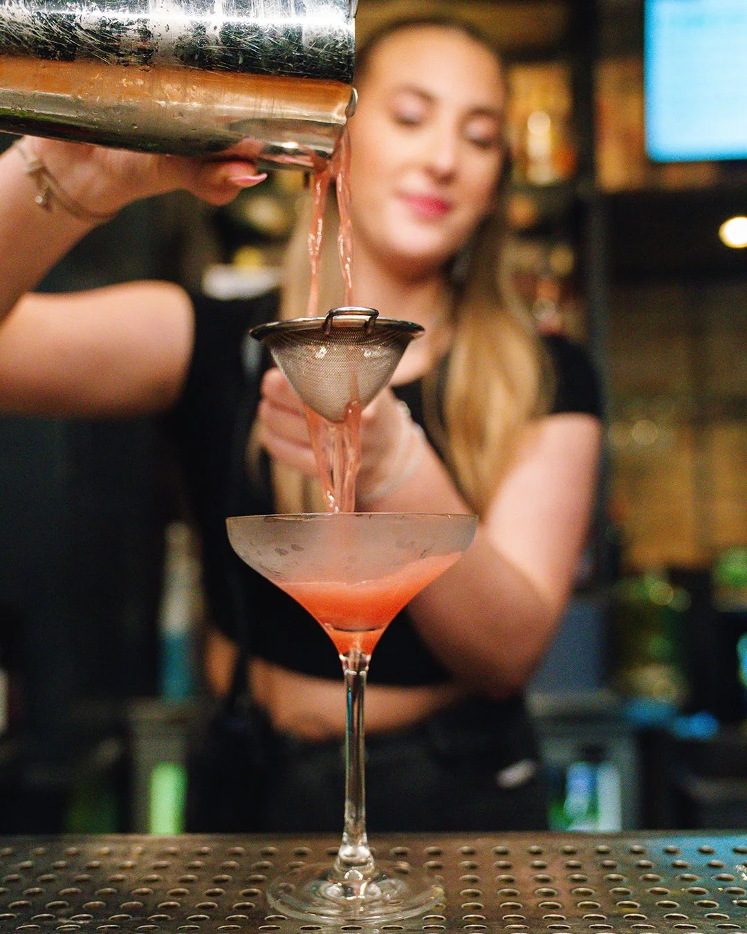 A bartender pours a pink cocktail from a shaker through a strainer into a martini glass in a bar.