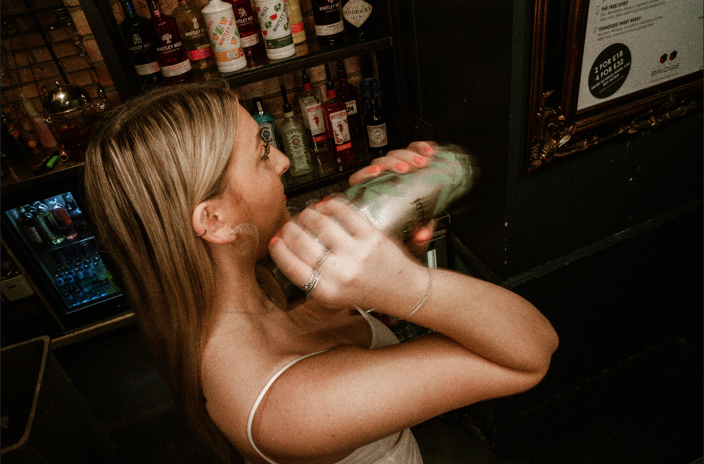 A woman with long blonde hair wearing earrings and a white spaghetti strap top, holding a cocktail shaker, behind a bar with liquor bottles.