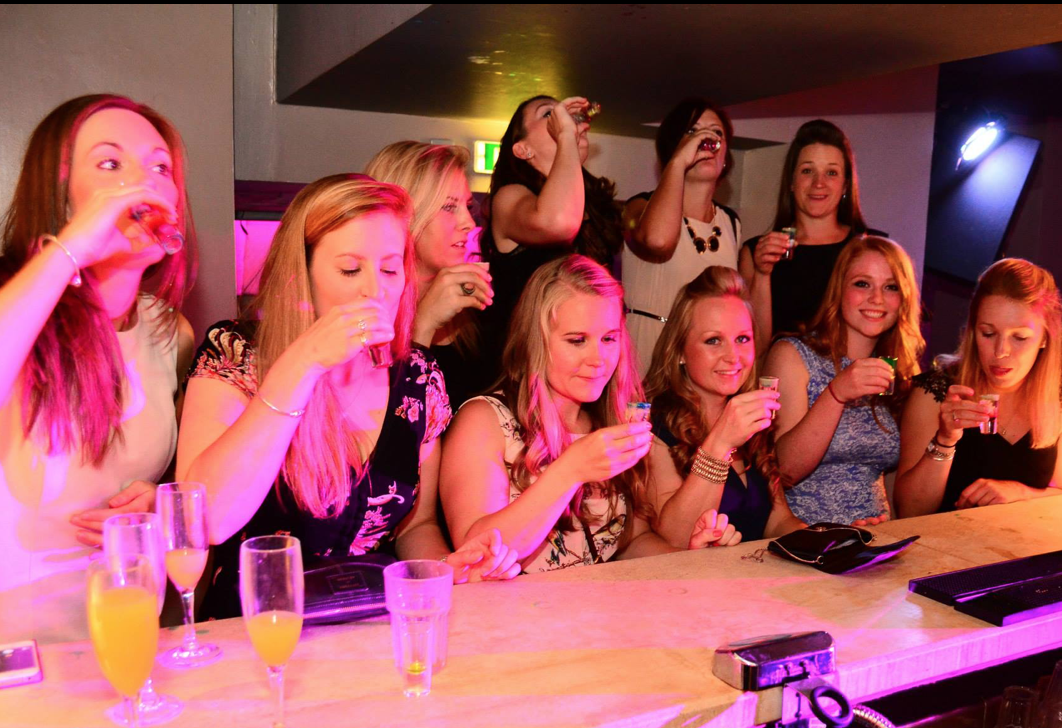 Group of ten women in a bar or club, some taking shots and others holding drinks, with pink and purple lighting.