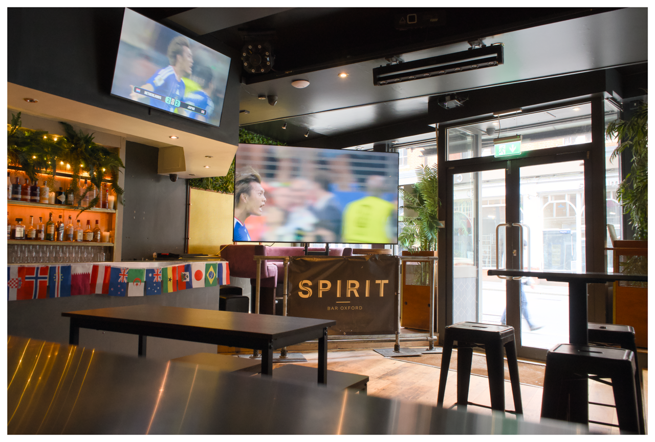 Interior of a bar or cafe with a large TV screen showing a soccer match, a bar shelf decorated with holiday greenery and flags from various countries, and a glass door entrance with plants inside.