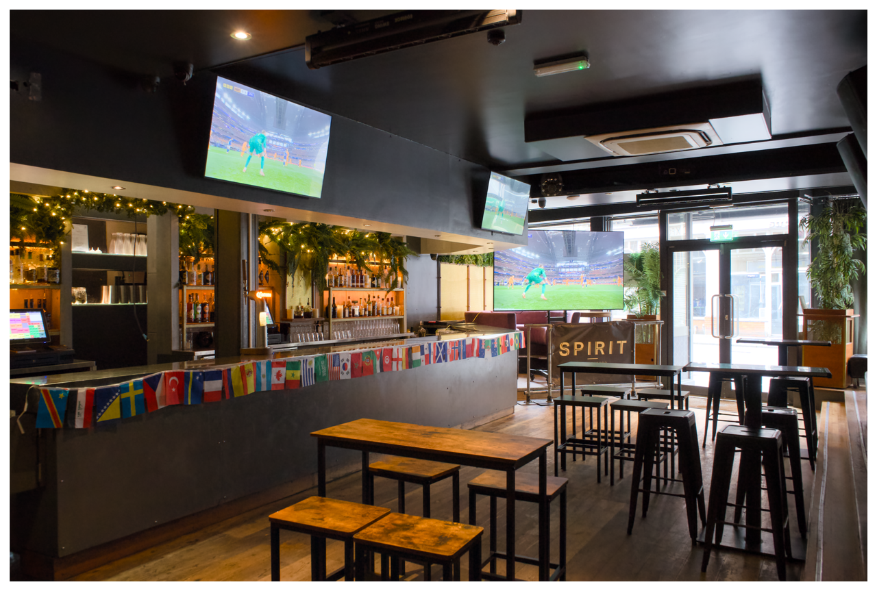 Interior of a sports bar with multiple flat-screen TVs showing a football game, a bar counter decorated with international flags, high tables and stools, and large windows with plants and natural light.