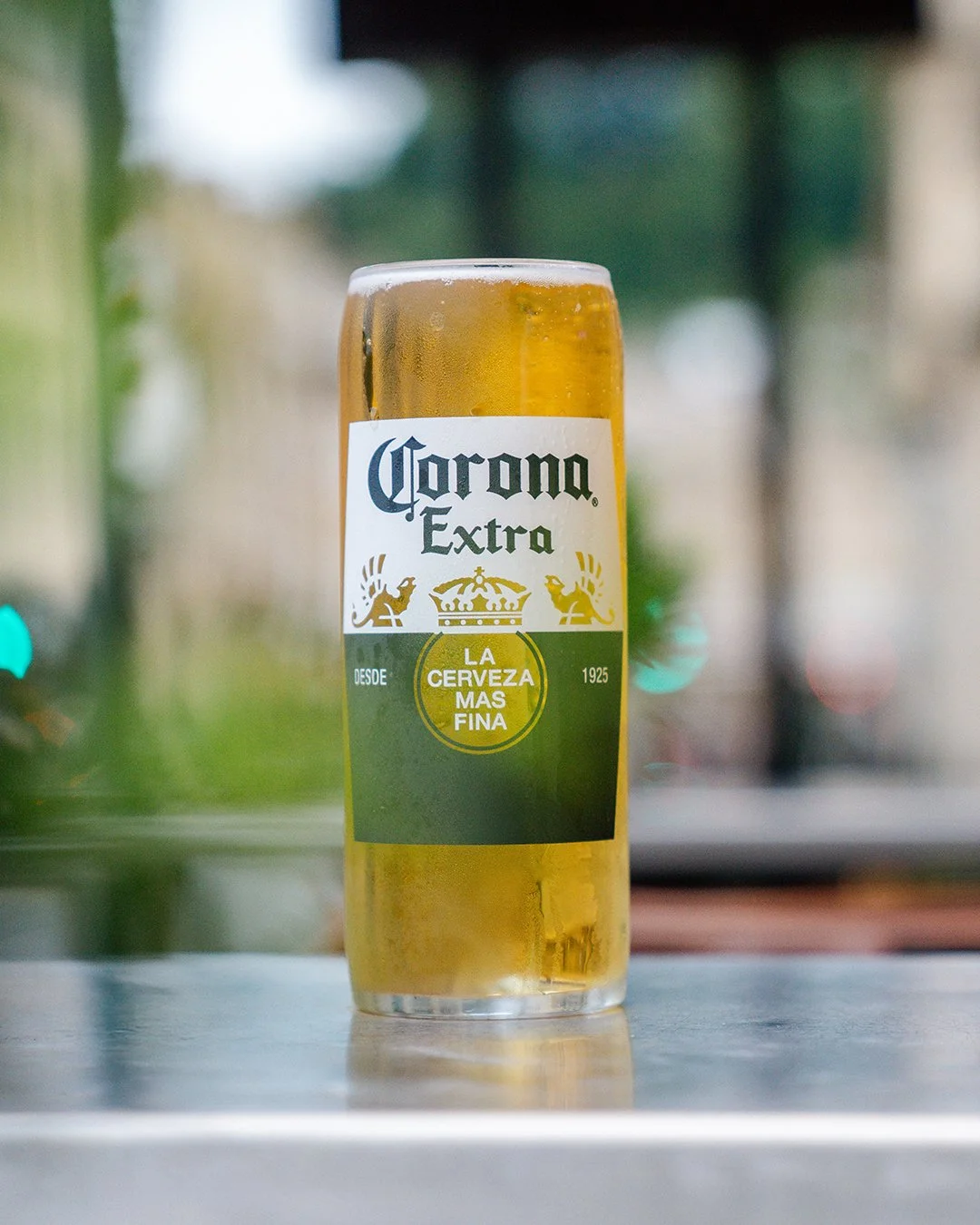 A glass of Corona Extra beer on a table with a blurred outdoor background.
