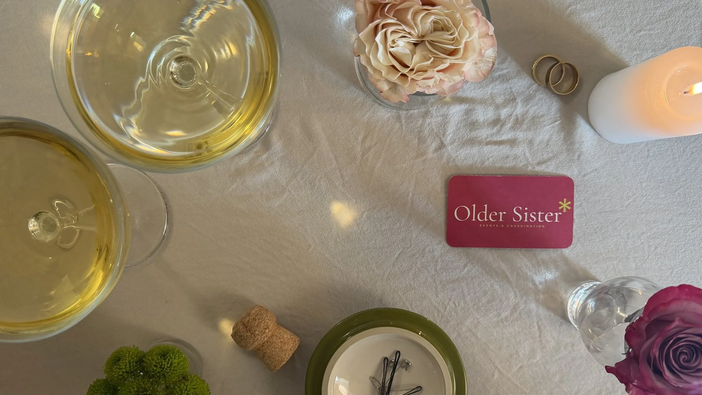 Older Sister Events business card on a cream linen table with champagne coupes, garden roses, a lit candle and a dish of safety pins
