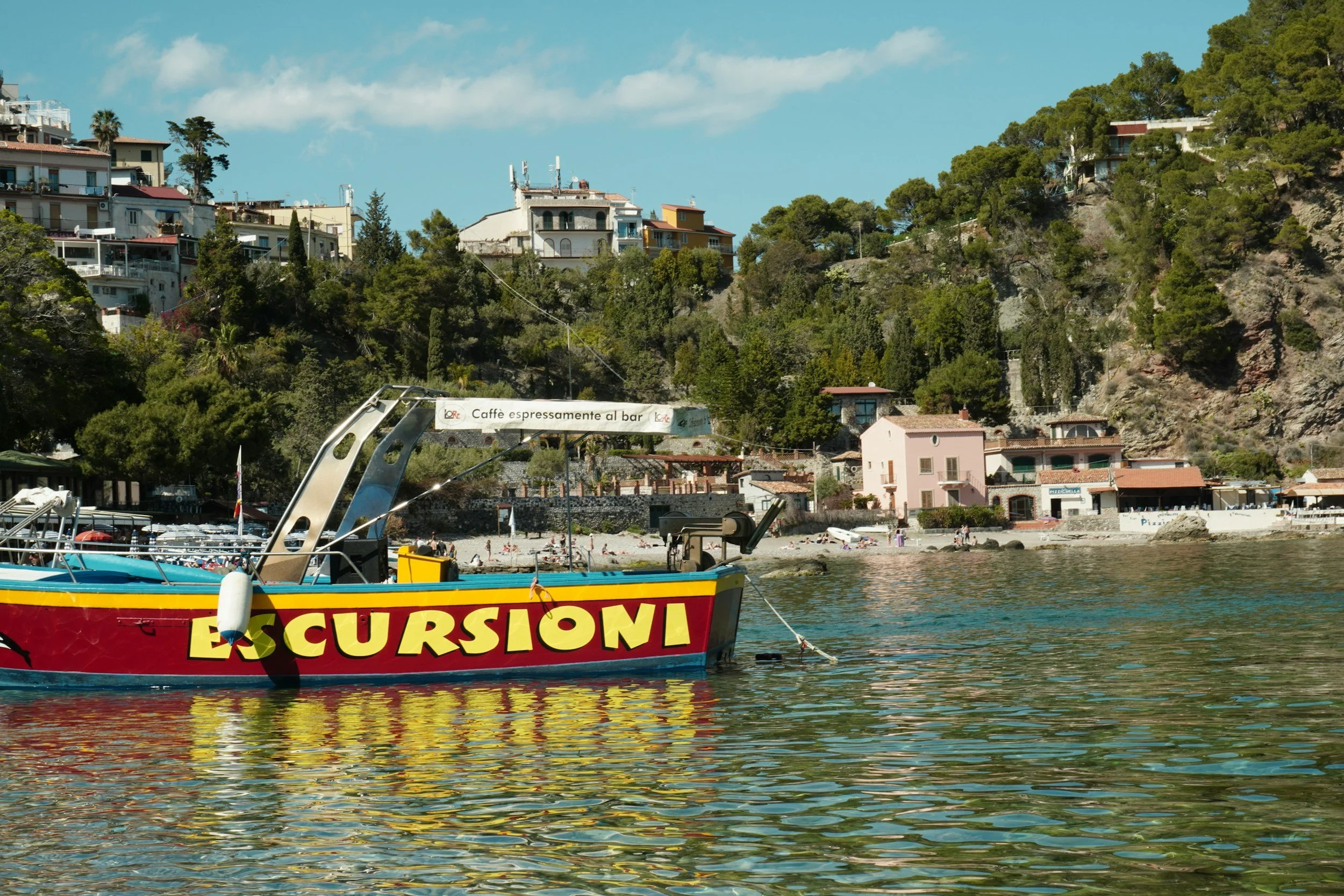 Colorful boat with 'Escursioni' written on it floating on water, with a beach and hillside houses in the background under a partly cloudy sky.