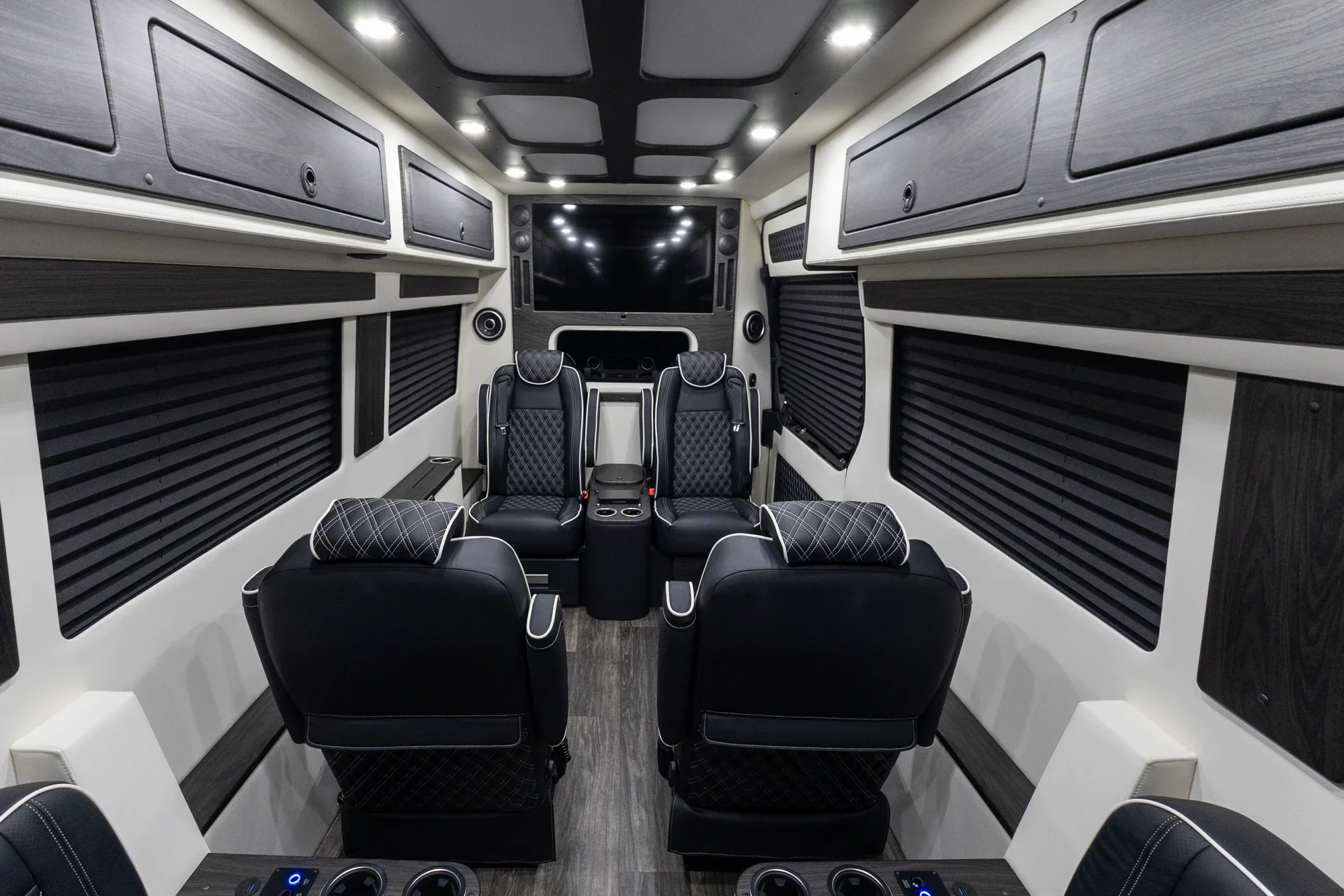 Interior of a luxury van or RV with black leather seats, multiple overhead storage compartments, a large flat-screen TV on the front wall, and sound speakers on each side.